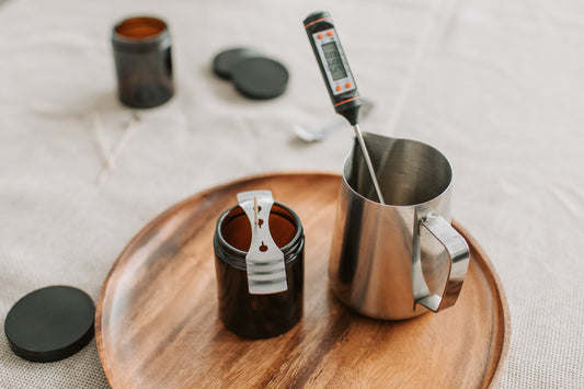 candle making supplies like a thermometer, pouring pitcher, and wick stabilizer on a wooden plate